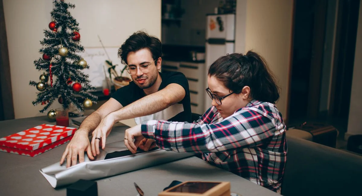 A couple enjoying togetherness while wrapping Christmas presents in a cozy indoor setting.